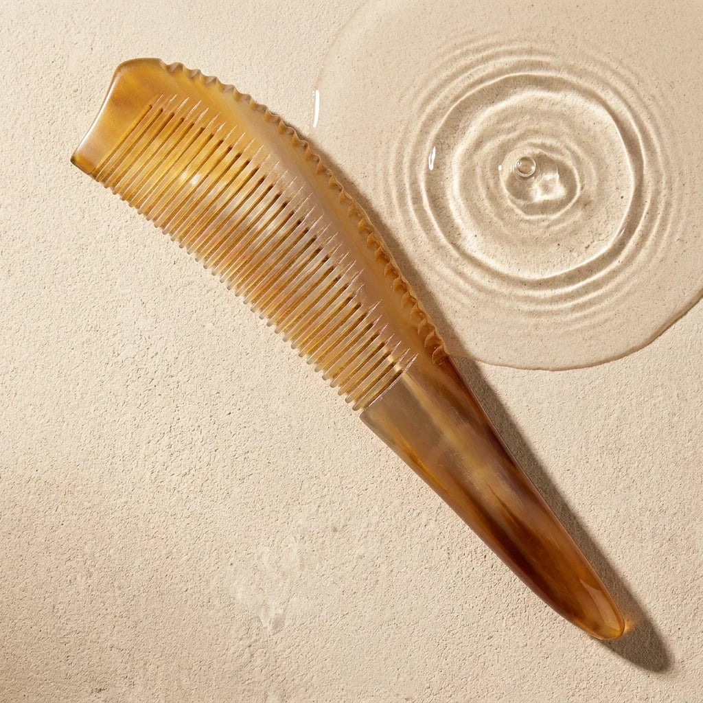 Pointed yellow horn comb resting beside subtle water ripples on a stone surface, suggesting a calm scalp ritual.