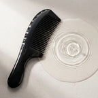 Black buffalo handled horn comb resting beside subtle water ripples on stone, suggesting a calm scalp ritual.