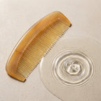 Translucent sheep horn comb resting beside delicate water ripples on a light stone surface, suggesting a calm scalp ritual.