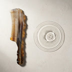 Striated yak horn comb beside water ripples on a light stone surface, suggesting a calm scalp ritual.