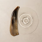 Curved natural horn comb beside subtle water ripples on a light surface, suggesting a calm scalp ritual.